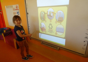 Dziewczynka stoi pod tablicą interaktywną i opisuje ilustrację przedstawiającą drogę recyklingu zużytego szkła.